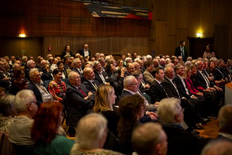 Aufnahme von Besucherinnen und Besuchern in einer Halle, die Sitzreihen sind eng bestuhlt, die Gäste blicken auf die Bühne.
