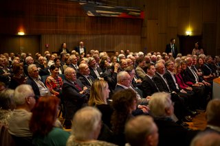 Aufnahme von Besucherinnen und Besuchern in einer Halle, die Sitzreihen sind eng bestuhlt, die Gäste blicken auf die Bühne.