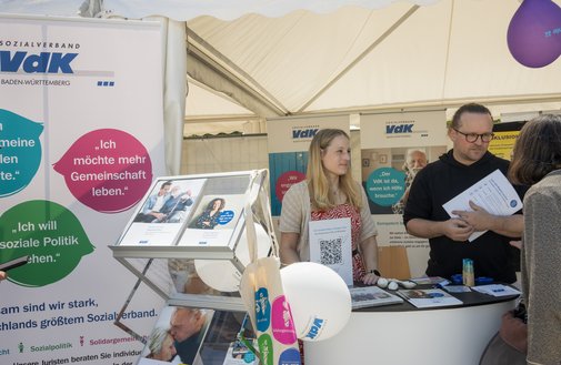 Freiburg Aktionstag Inklusion 2025 Beratung Blick von vorne auf den VdK-Infostand beim Aktionstag Inklusion in Freiburg, hinter einer kleinen Theke stehen zwei Mitarbeiter im Gespräch mit einer Ratsuchenden, die von hinten zu sehen ist. Am Stand steht noch ein Prospektständer mit mehreren Broschüren und Flyern,Luftballons wehen. Ein Roll-Up ist zu sehen,darauf verschiedene Slogans, zum Beispiel "Wir fordern soziale Rechte"