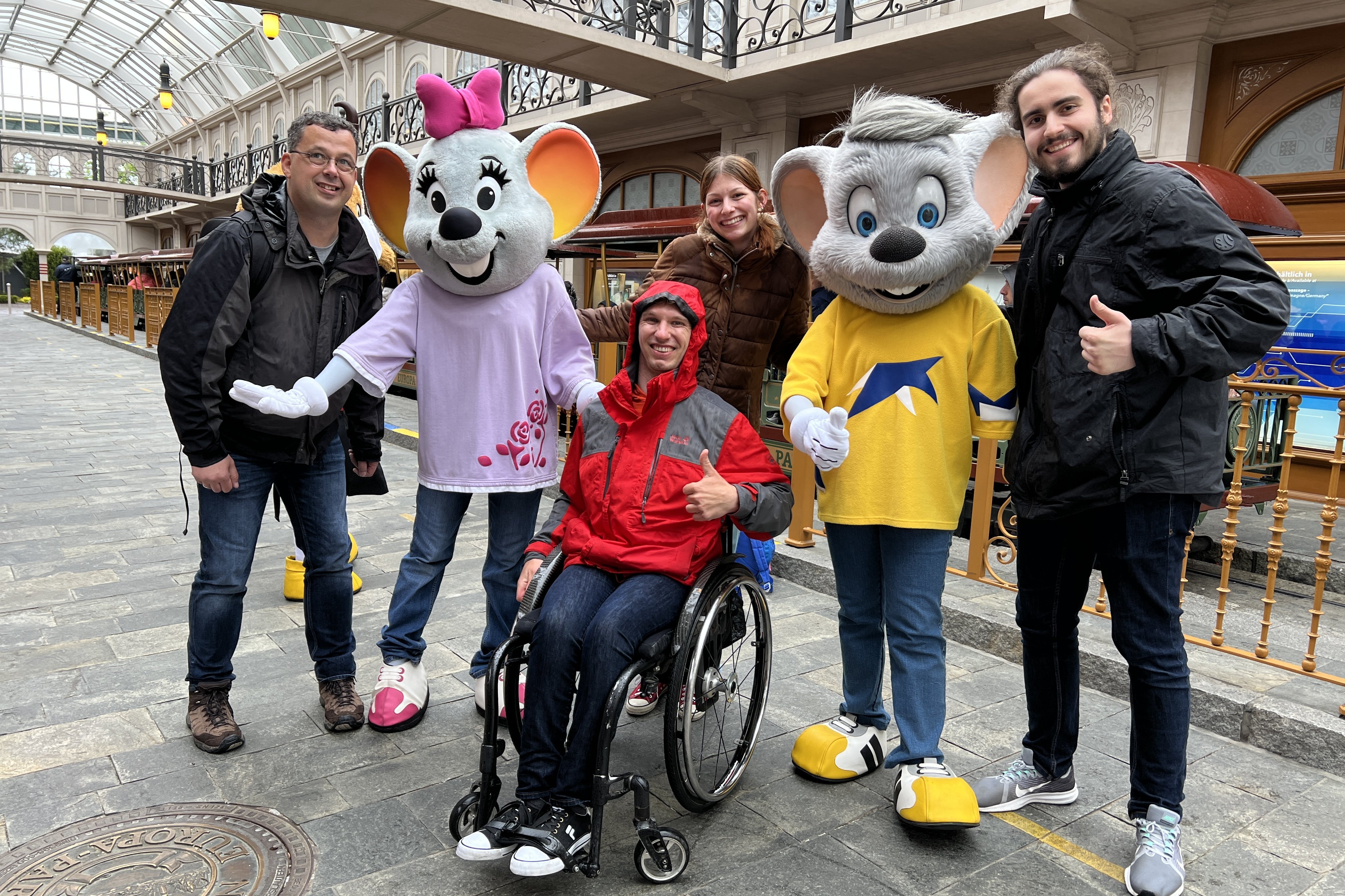 Das Team Barrierefreiheit von Freizeitpark Erlebnis posiert mit der Euro-Maus im Europa-Park, mittendrin ein Rollstuhlfahrer mit dem der Europa-Park live auf Barrierefreiheit getestet wird.