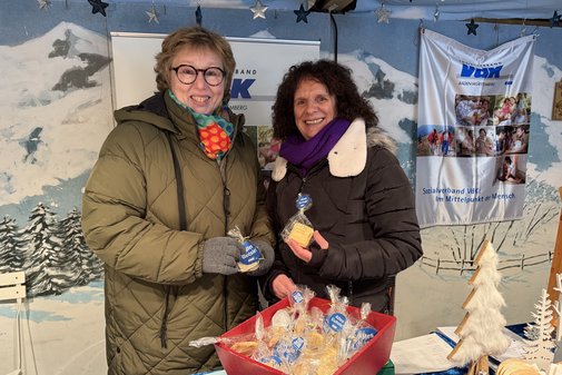 Zwei Frauen im VdK-Stand lachen in die Kamera,im Vordergrund sind die verpackten Solidarit&auml;tspl&auml;tzchen zu sehen.