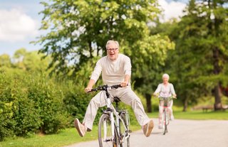 Älterer Mann sitzt fröhlich auf einem Fahrrad und fährt bei strahlendem Sonnenschein die Straße entlang