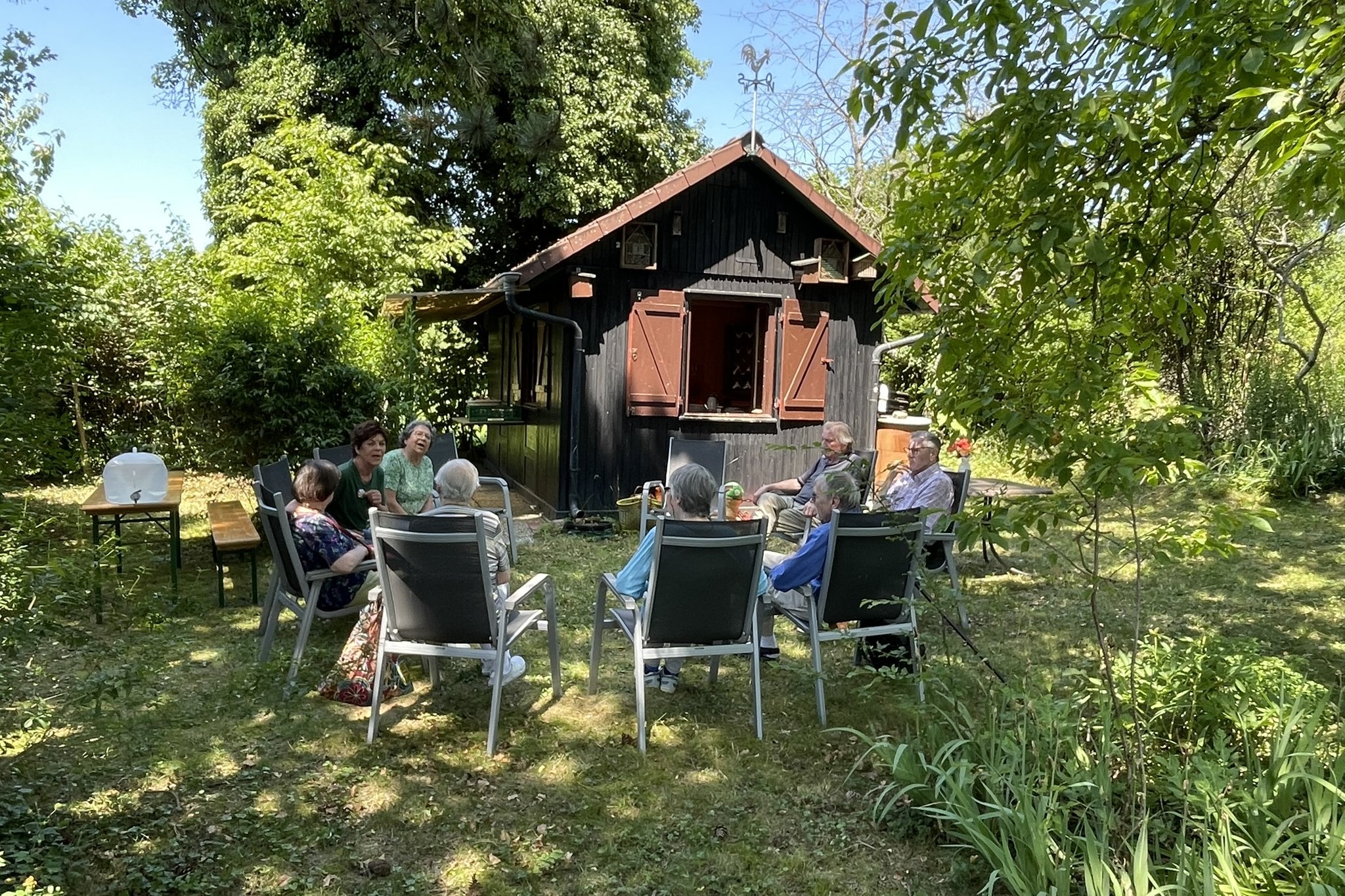 Gr&uuml;ner Garten mit einem Baumhaus, davor sitzen &auml;ltere Menschen in Gartenst&uuml;hlen im Kreis, ins Gespr&auml;ch vertieft. 