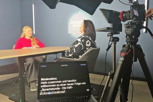 Dreharbeiten im Studio, am Tisch sitzen die Patientenberaterin und die Moderatorin ins Gespräch vertieft. Rechts im Bild ist eine Kamera zu sehen, die mitfilmt.