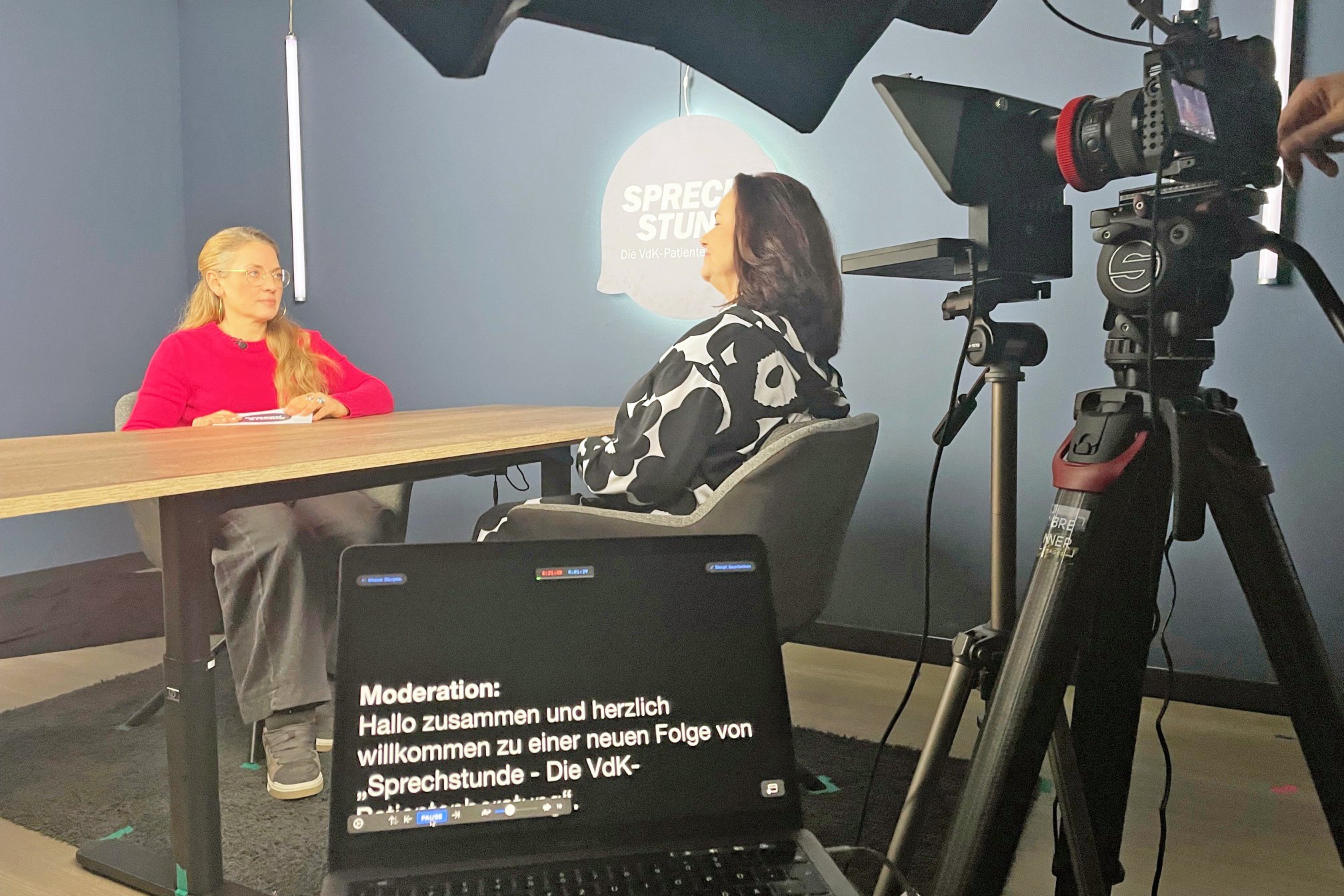 Videoformat Sprechstunde Patientenberatung Dreharbeiten Dreharbeiten im Studio, am Tisch sitzen die Patientenberaterin und die Moderatorin ins Gespräch vertieft. Rechts im Bild ist eine Kamera zu sehen, die mitfilmt.
