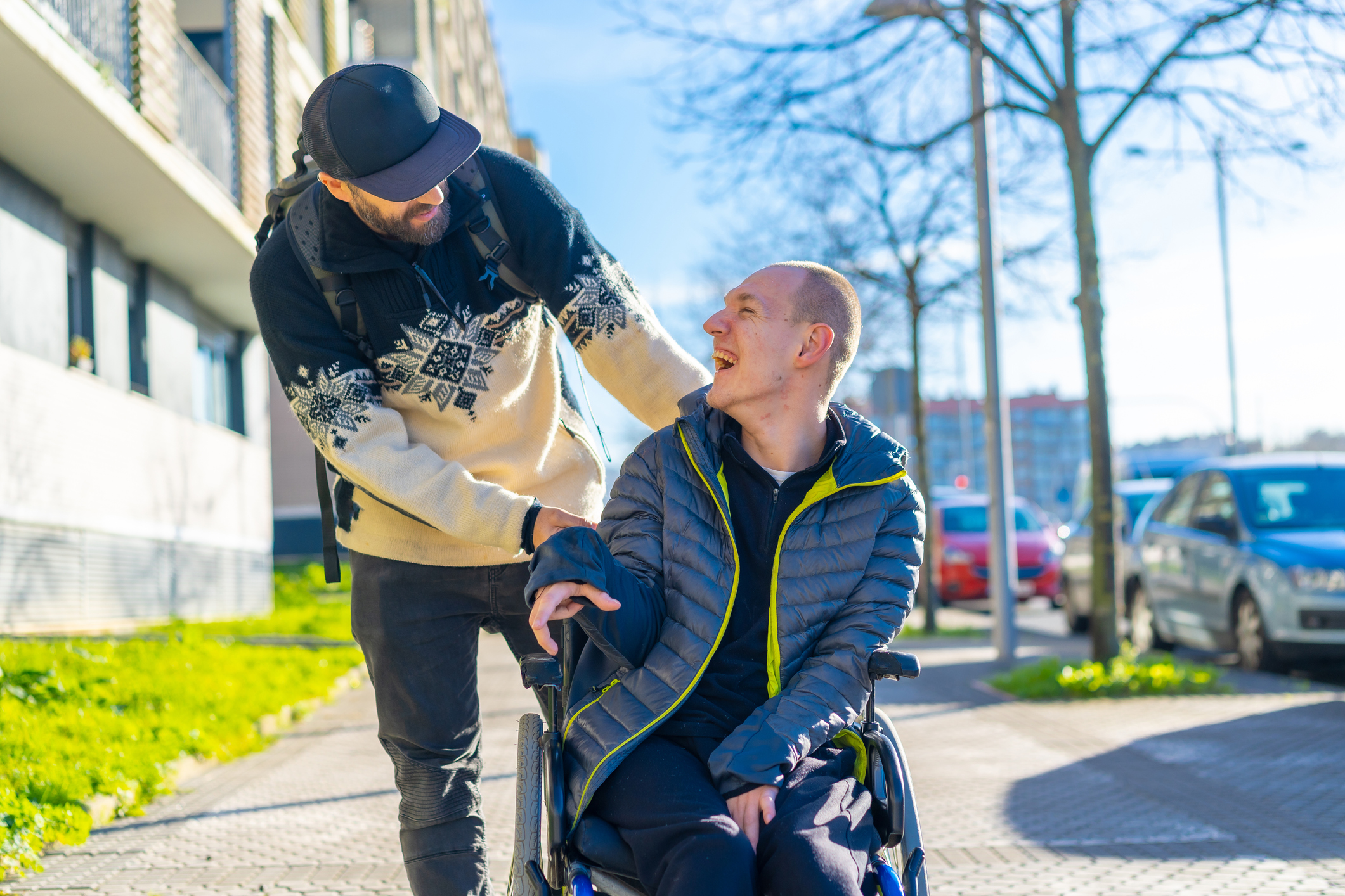 Eine behinderte Person im Rollstuhl wird von einem lächelnden Freund geschoben