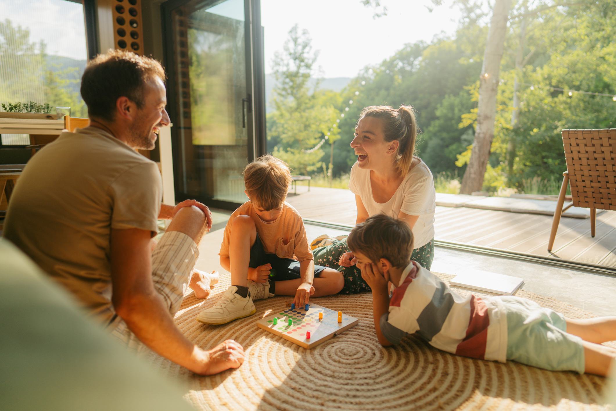 Rente DRV Tipps Kindererziehungszeiten Altersvorsorge Blick in ein Wohnzimmer, die Sonne scheint herein, auf dem Teppich sitzen Mutter und Vater mit ihren zwei Kindern und spielen mit ihnen lachend das Spiel "Mensch ärgere dich nicht".