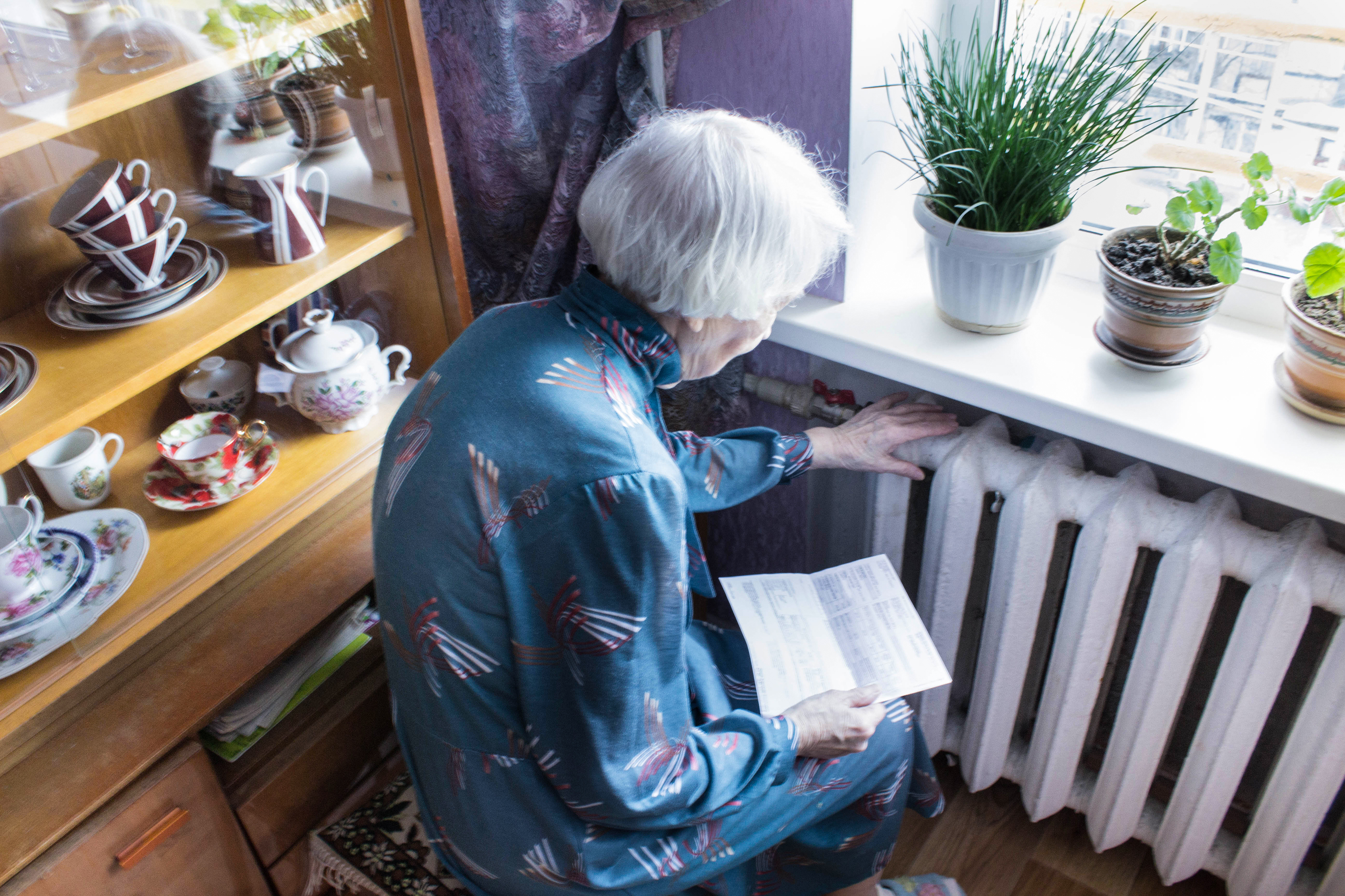Ältere Frau sitzt vor ihrem Heizkörper und wärmt daran eine Hand, in der anderen Hand hält sie die Gasrechnung