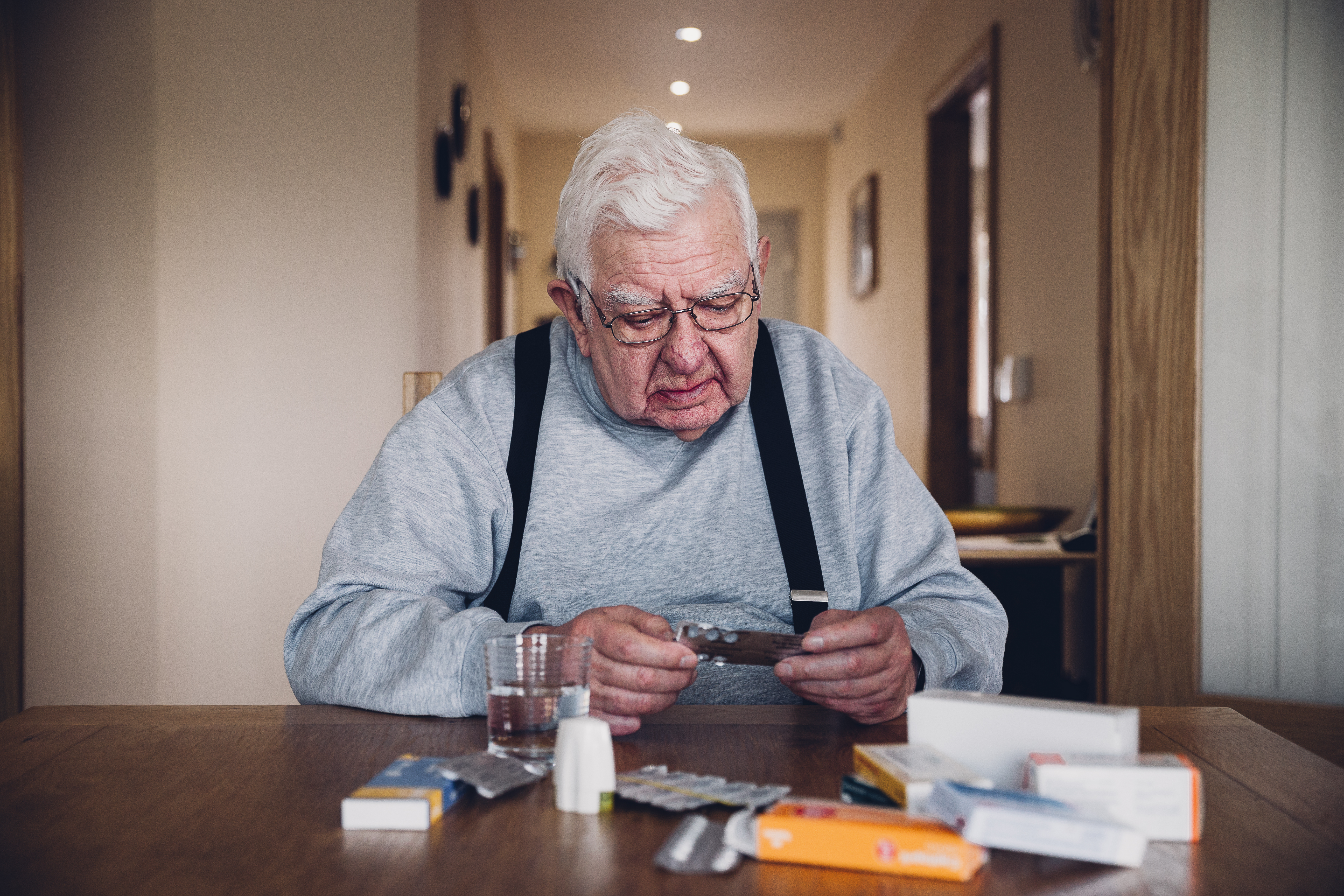 Medikamente Senior Rentner Management Älterer Mann sitzt am Tisch, er trägt eine Latzhose. Vor ihm liegen auf einem Tisch zahlreiche Tabletten, Tablettenverpackungen und leere Tablettendosen. Er liest angestrengt eine Packungsbeilage.