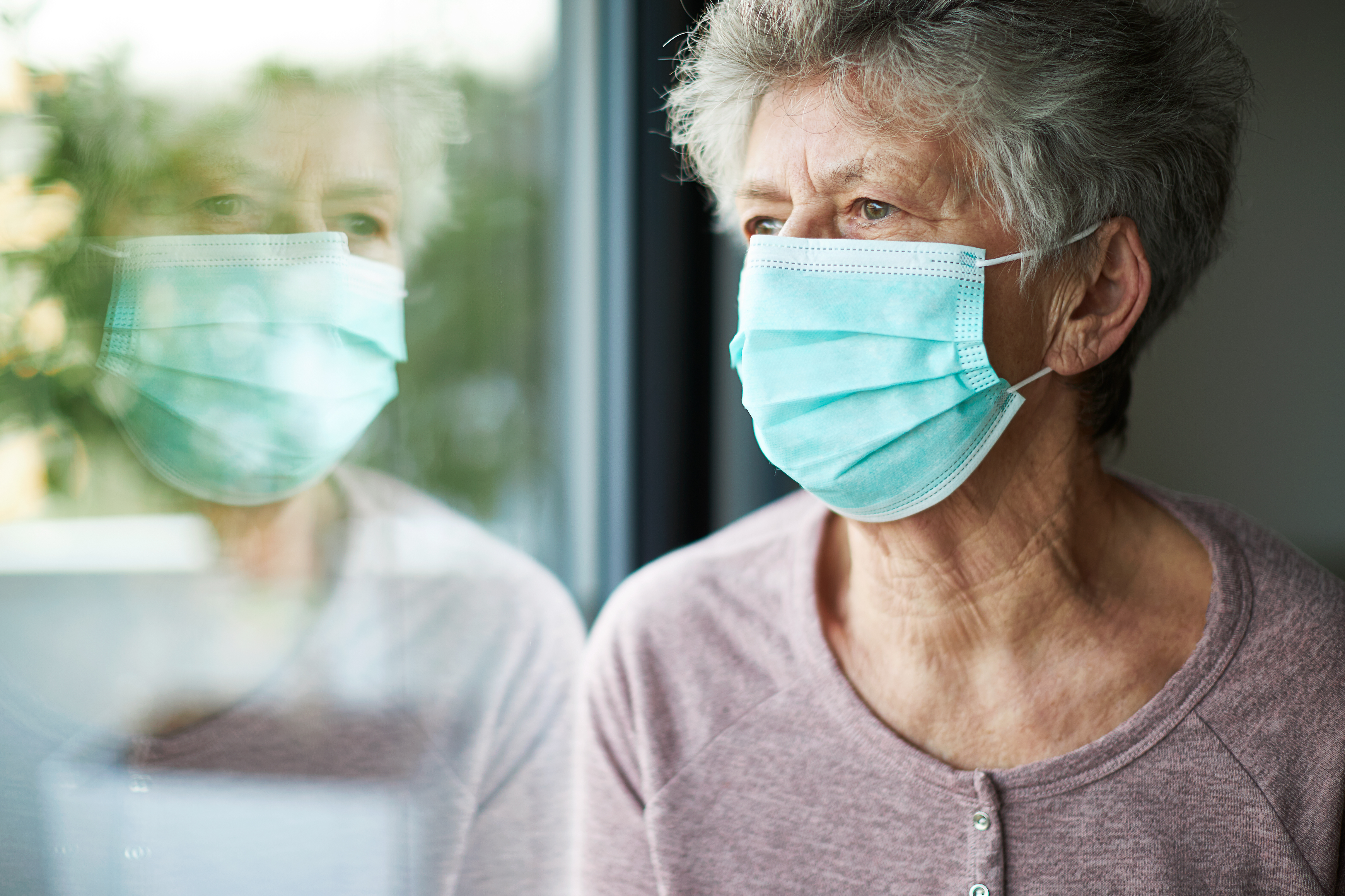 Eine alte Frau trägt eine OP-Maske und schaut aus dem Fenster