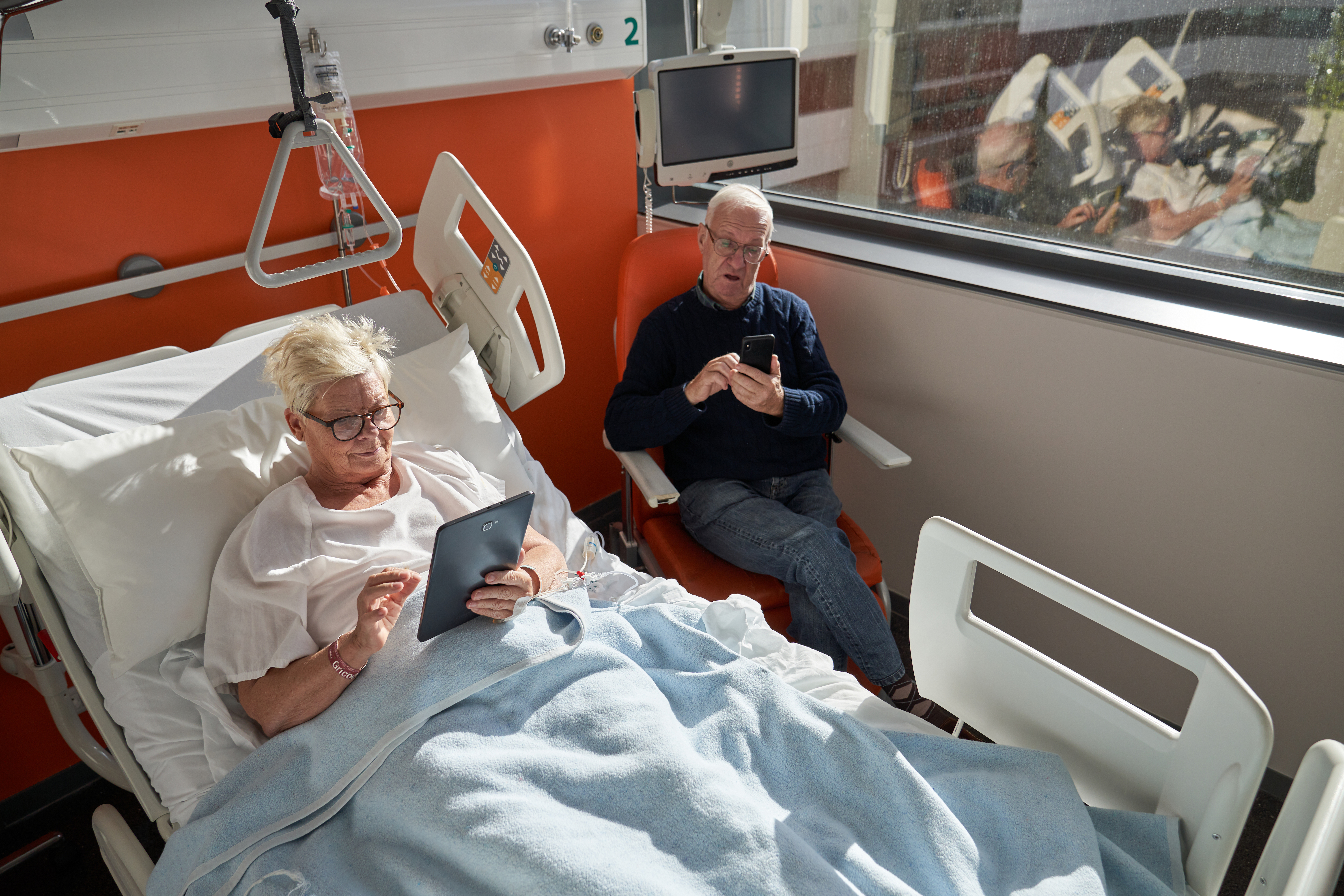 &Auml;lterer Mann sitzt neben seiner Frau in einem Stuhl im Krankenhaus, die Frau liegt in einem Krankenhauskittel in einem Krankenhausbett und bedient ein Tablet.
