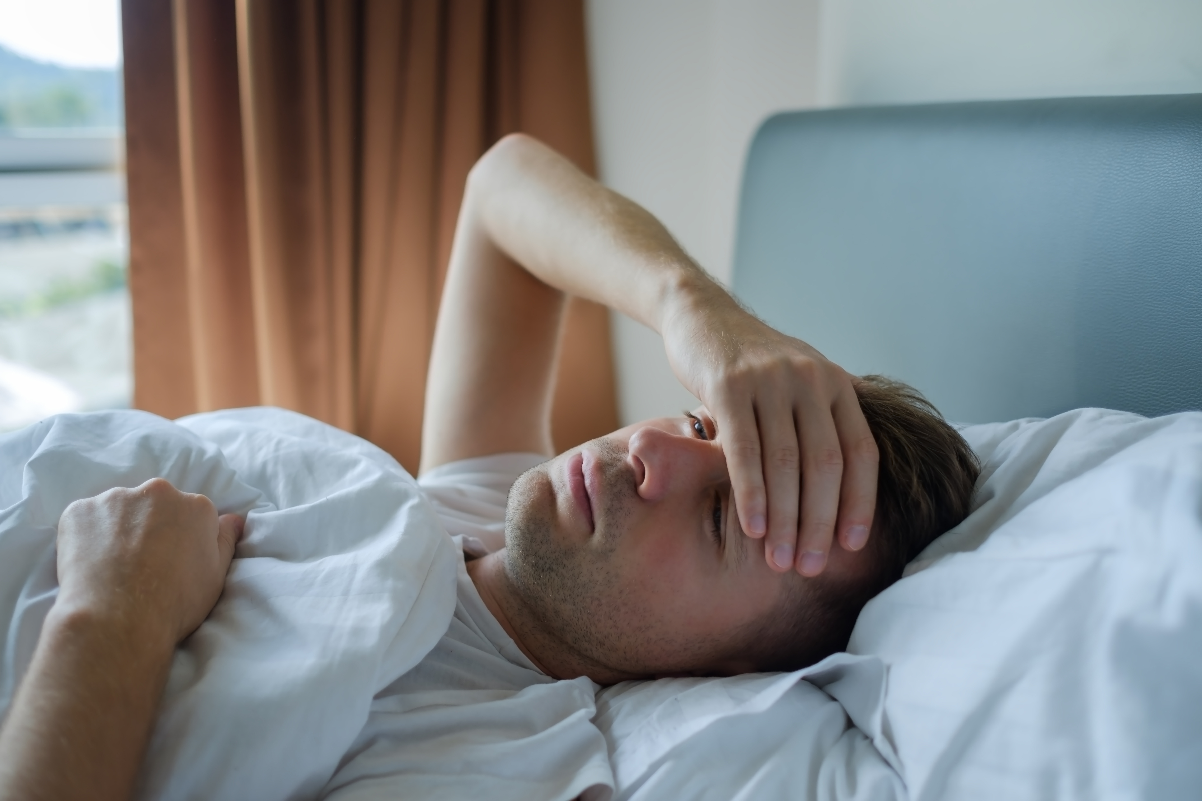 Mann liegt krank zu Hause in seinem Bett, die Bettdecke ist bis zur Brust hochgezogen, mit einer Hand greift er sich an die Stirn, um die Temperatur zu fühlen.