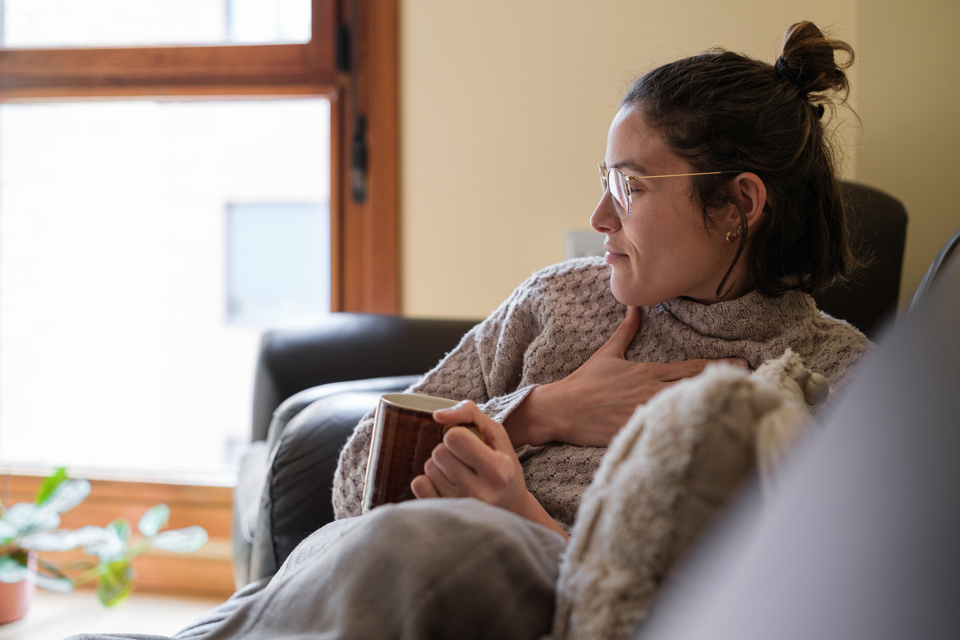 Frau greift sich wegen Halsschmerzen an den Hals, sie sitzt dabei auf dem Sofa, in der anderen Hand hält sie eine Tasse Tee