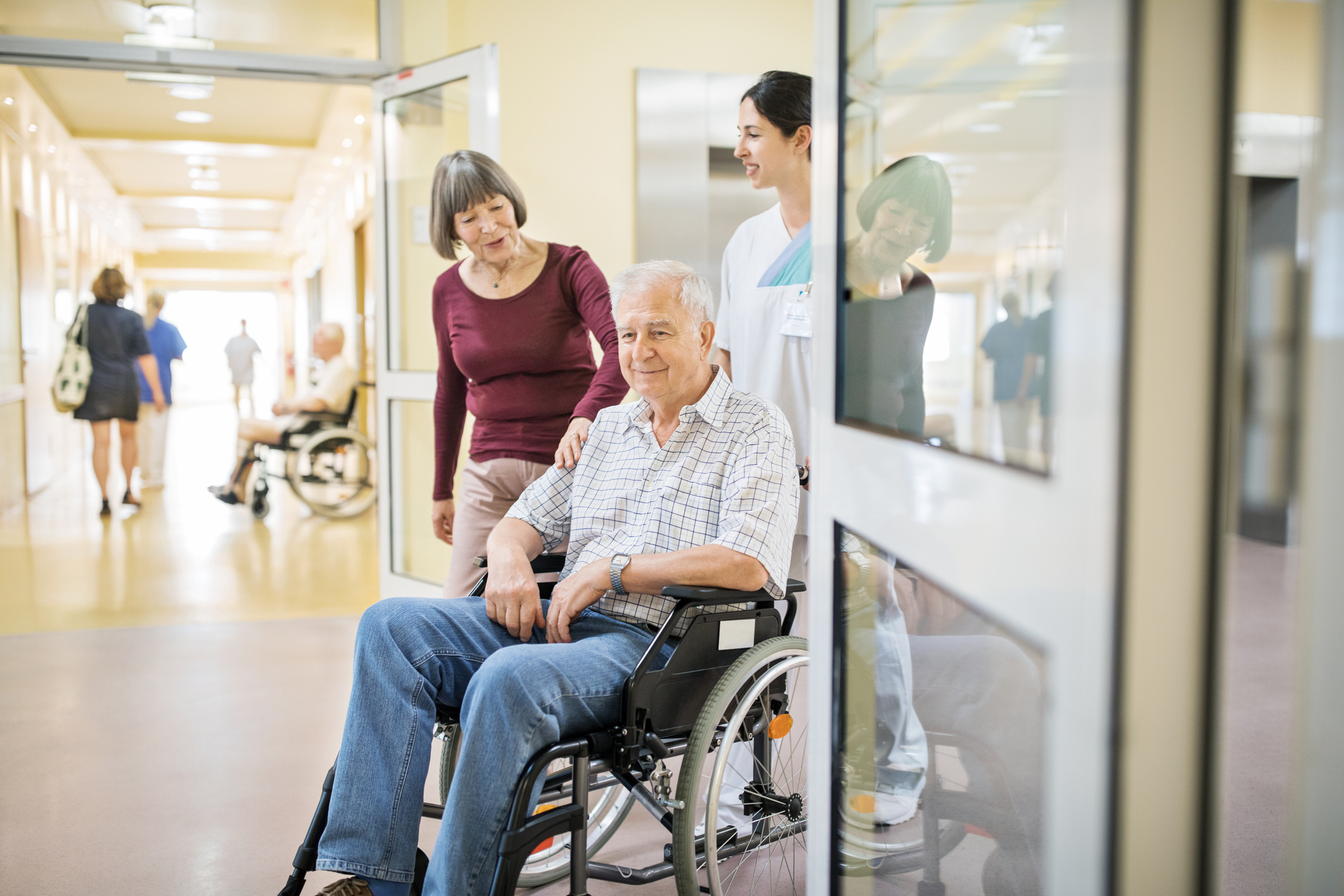 Älterer Mann, der im Rollstuhl sitzt und mit Frau und Krankenschwester durch den Krankenhauskorridor geht.