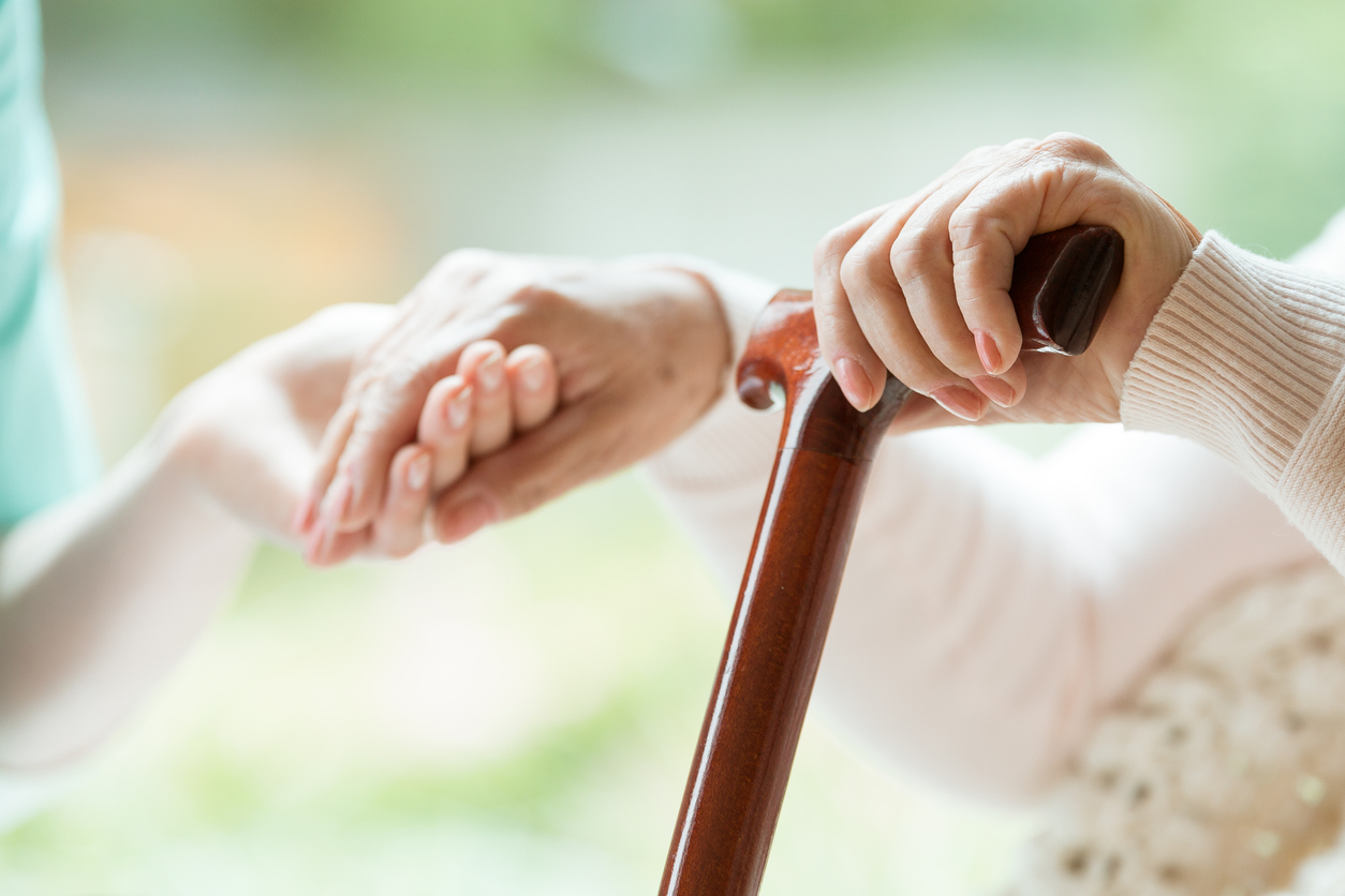 Ältere Person stützt sich auf einen Gehstock, Pflegekraft greift die andere Hand um beim Aufstehen zu helfen