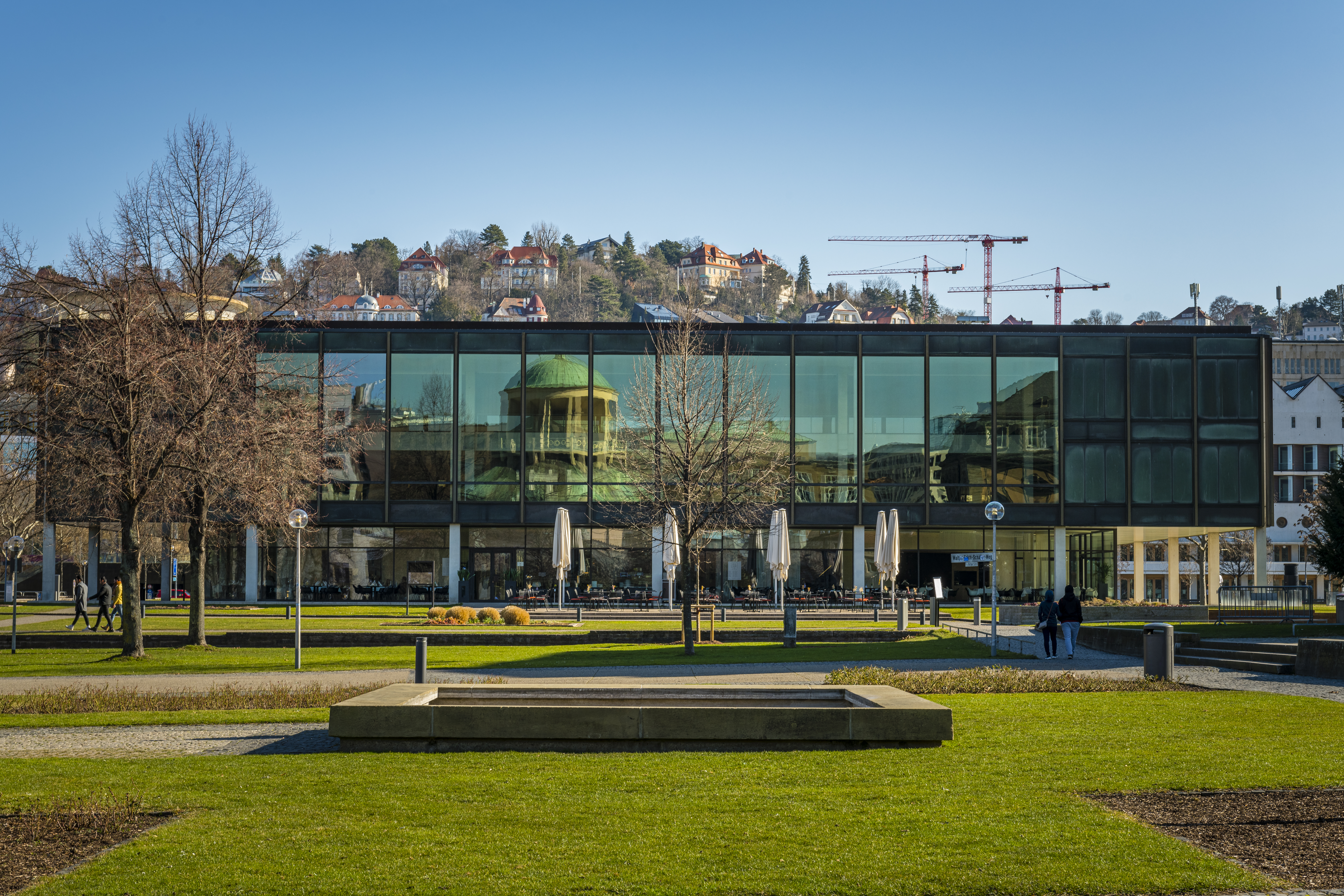 Au&szlig;enansicht des Landtags Baden-w&uuml;rttemberg in Stuttgart: Ein gl&auml;senernes Geb&auml;ude im Hintergrund, in dem sich ein gegen&uuml;berliegendes Geb&auml;ude mit einer Kuppel spiegelt. Vor dem Geb&auml;ude eine gr&uuml;ne Wiese und ein Brunnen. Im Hintergrund ist die Spitze eines Berges zu erkennen mit zahlreichen H&auml;usern.