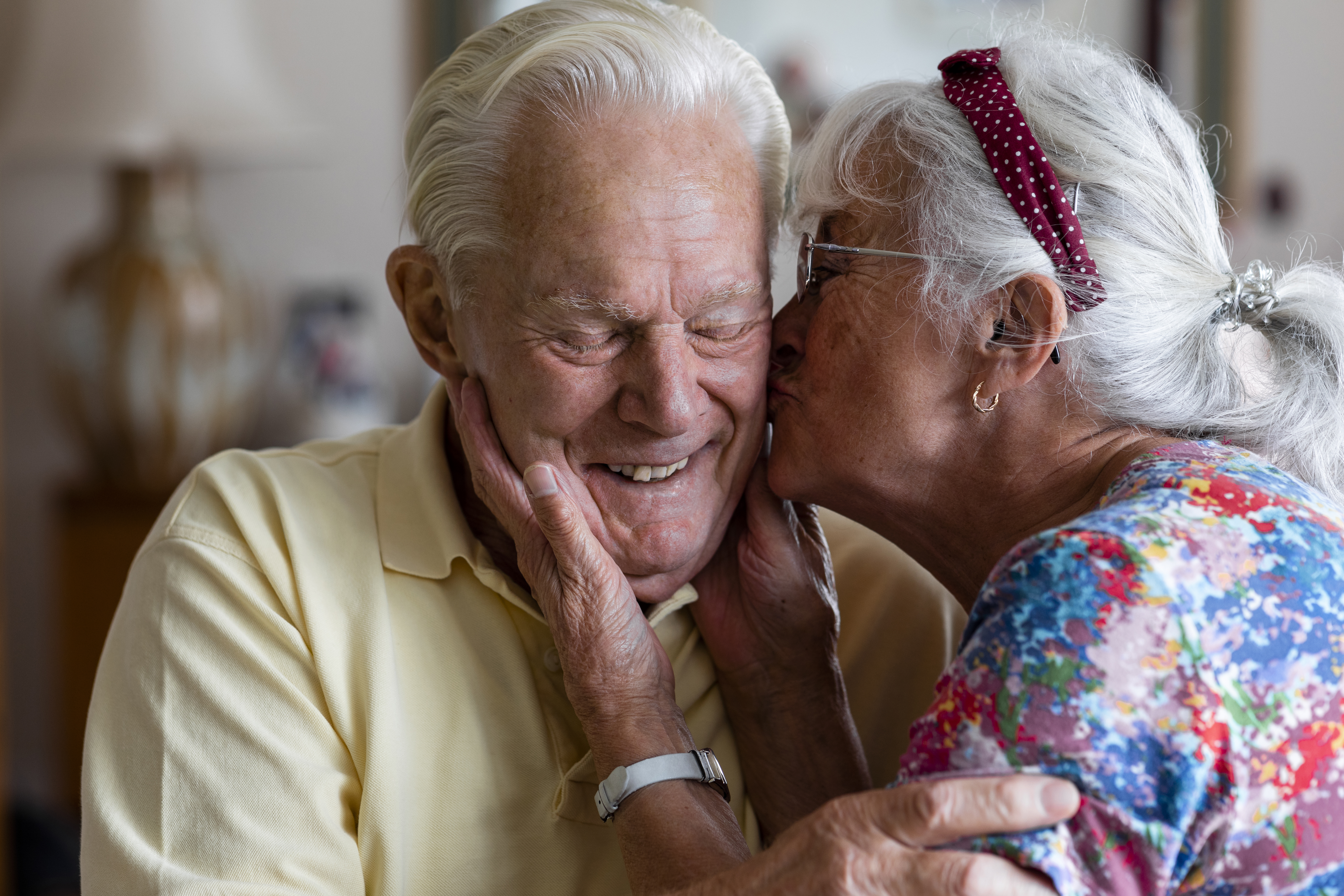 Senior-Paar zu Hause: die Frau küsst die Wange ihres Mannes während er mit geschlossenen Augen lacht