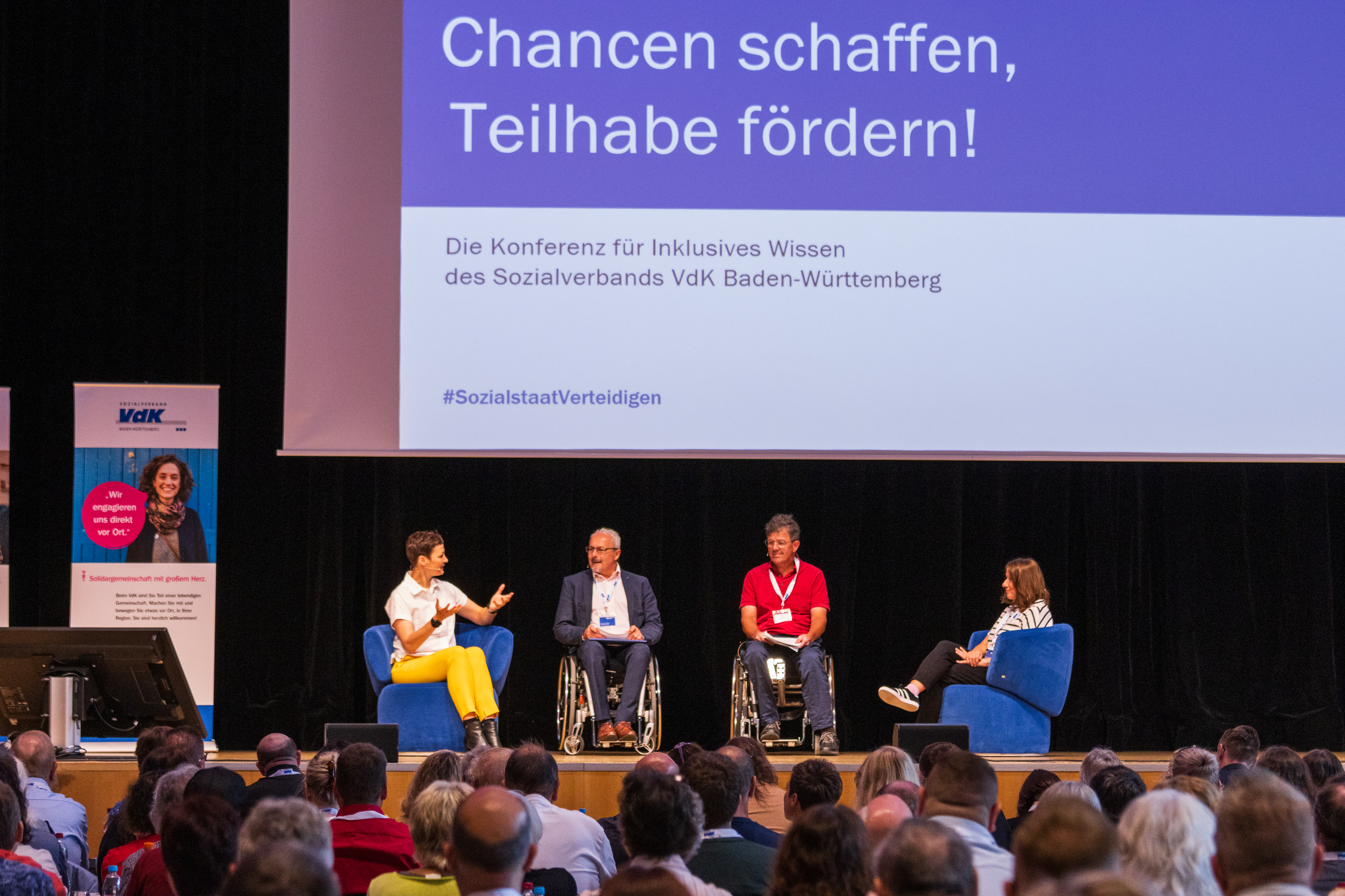 SBV-Konferenz 2025 Podiumsgespräch Barrierefreiheit Inklusion Teilhabe Blick auf die Bühne vom Publikum aus, zu sehen sind vier Personen, die auf Stühlen auf der Bühne sitzen und ins Gespräch vertieft sind, im Hintergrund ist eine Leinwand zu sehen auf der steht "Chancen schaffen, Teilhabe fördern!"