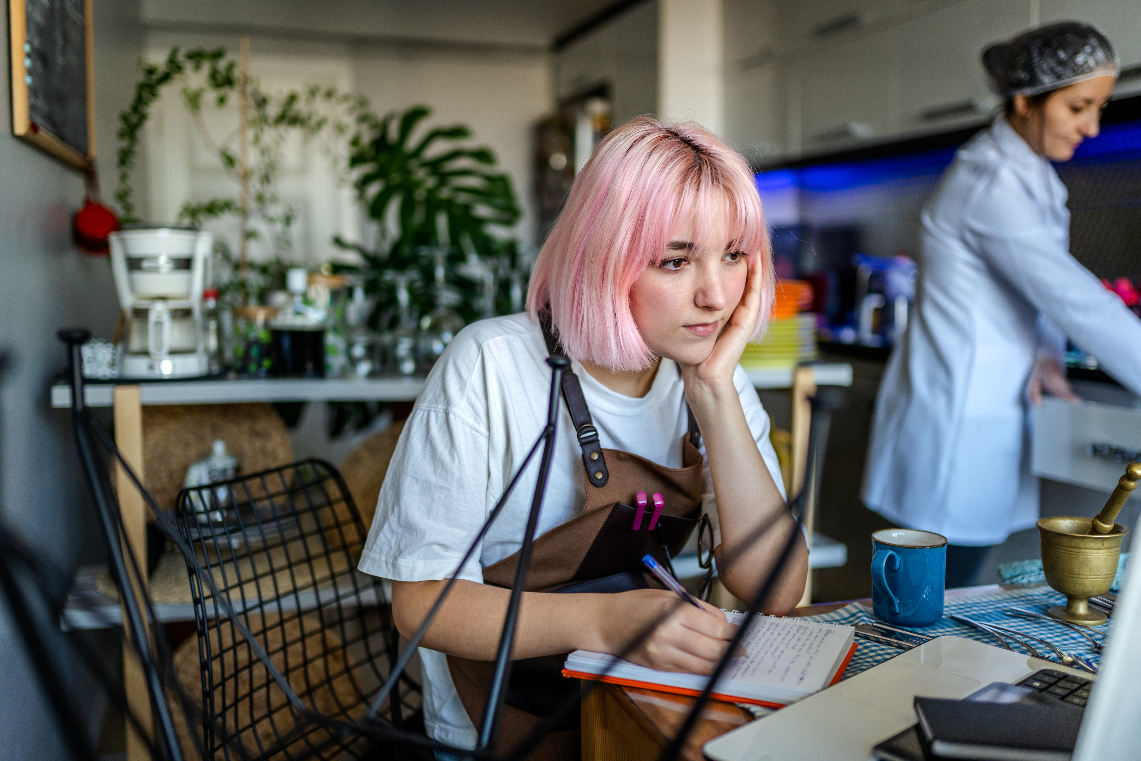 Studentin arbeitet in einem Caf&eacute; als Minijob, sie steht an der Theke und blickt, auf ihren Ellbogen gesch&uuml;tzt, auf die Kasse, rechts von ihr im Hintergrund steht eine Kollegin.