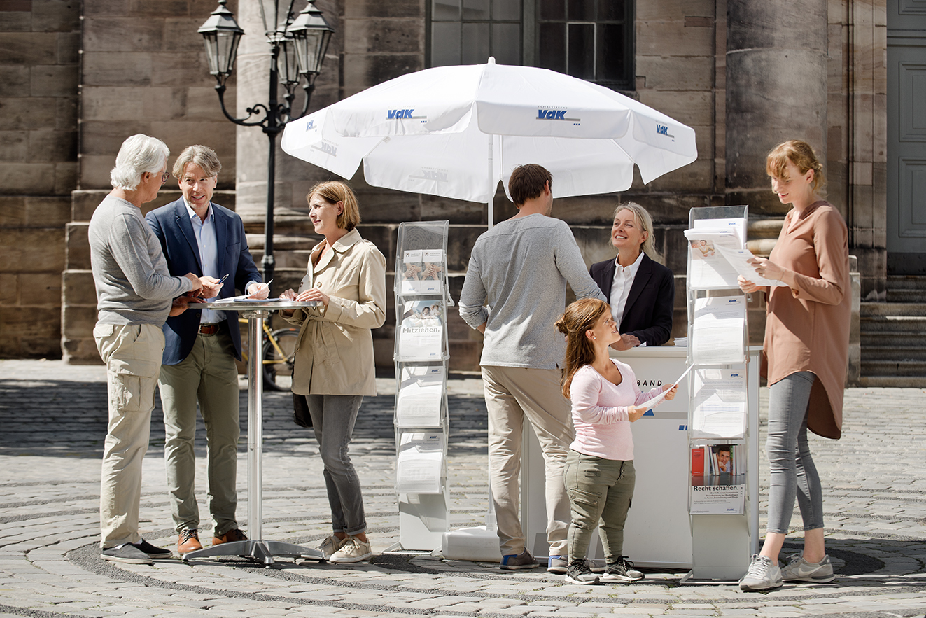 Ein VdK-Infostand in einer Fu&szlig;g&auml;ngerzone, eine Frau spricht mit einem Passanten, am Stehtisch daneben unterhalten sich Menschen. 