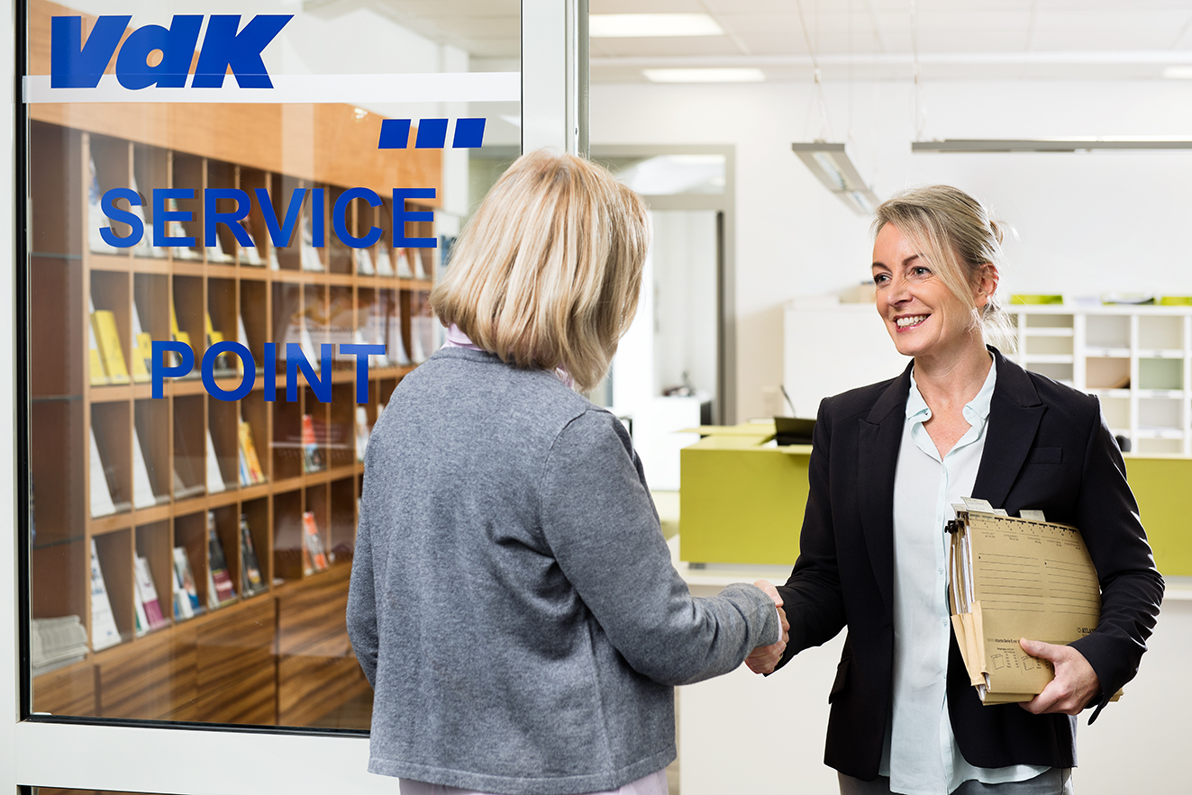 Eine VdK-Mitarbeiterin begr&uuml;&szlig;t eine Frau an der T&uuml;r einer VdK-Beratungsstelle. Sie hat eine Aktenmappe unter dem Arm. 