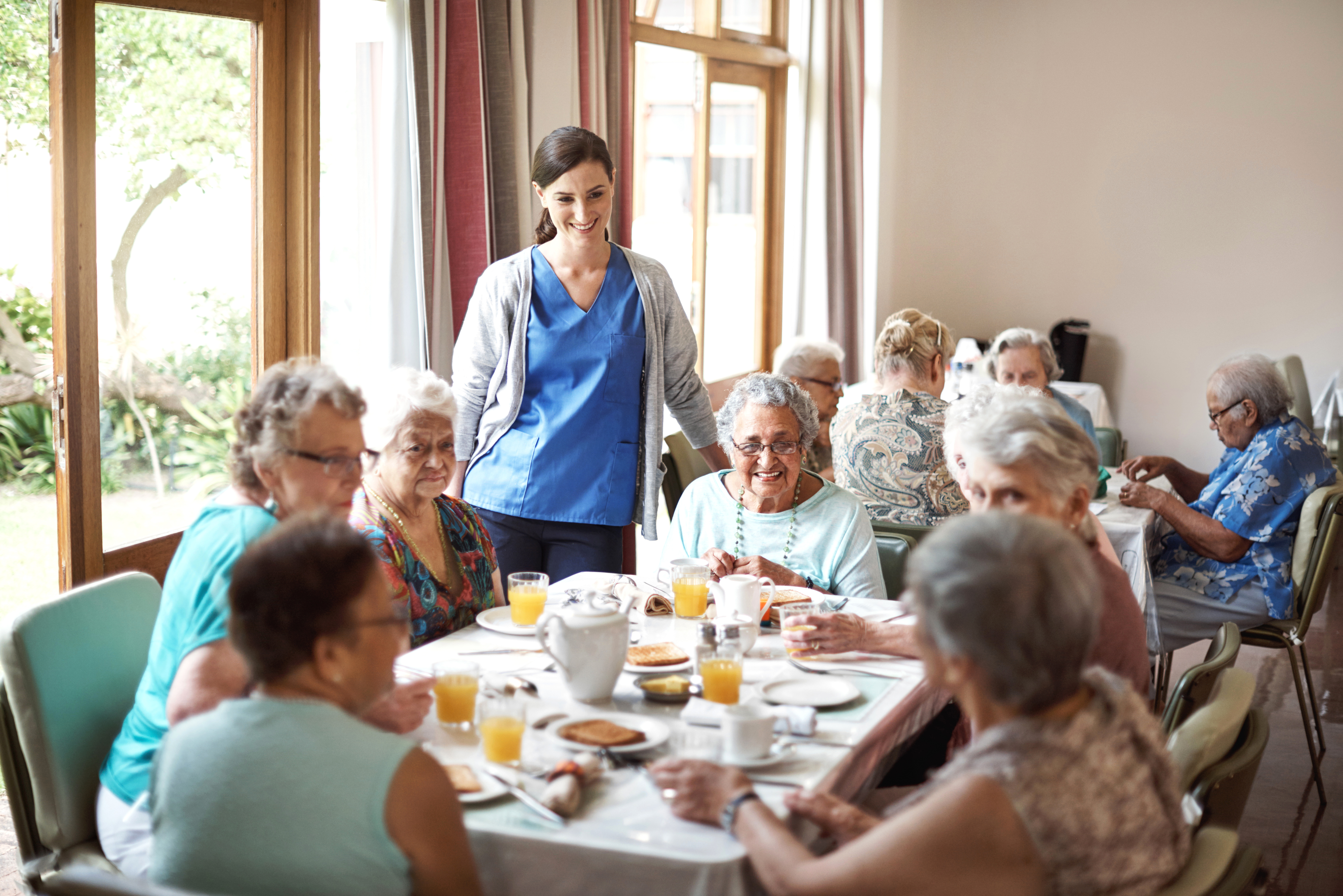 Esstisch in einem Seniorenheim, viele &auml;ltere Menschen sitzen zusammen am Tisch, unterhalten sich und essen. Am Tisch steht eine Pflegerin und erz&auml;hlt etwas oder h&ouml;rt gerade zu.