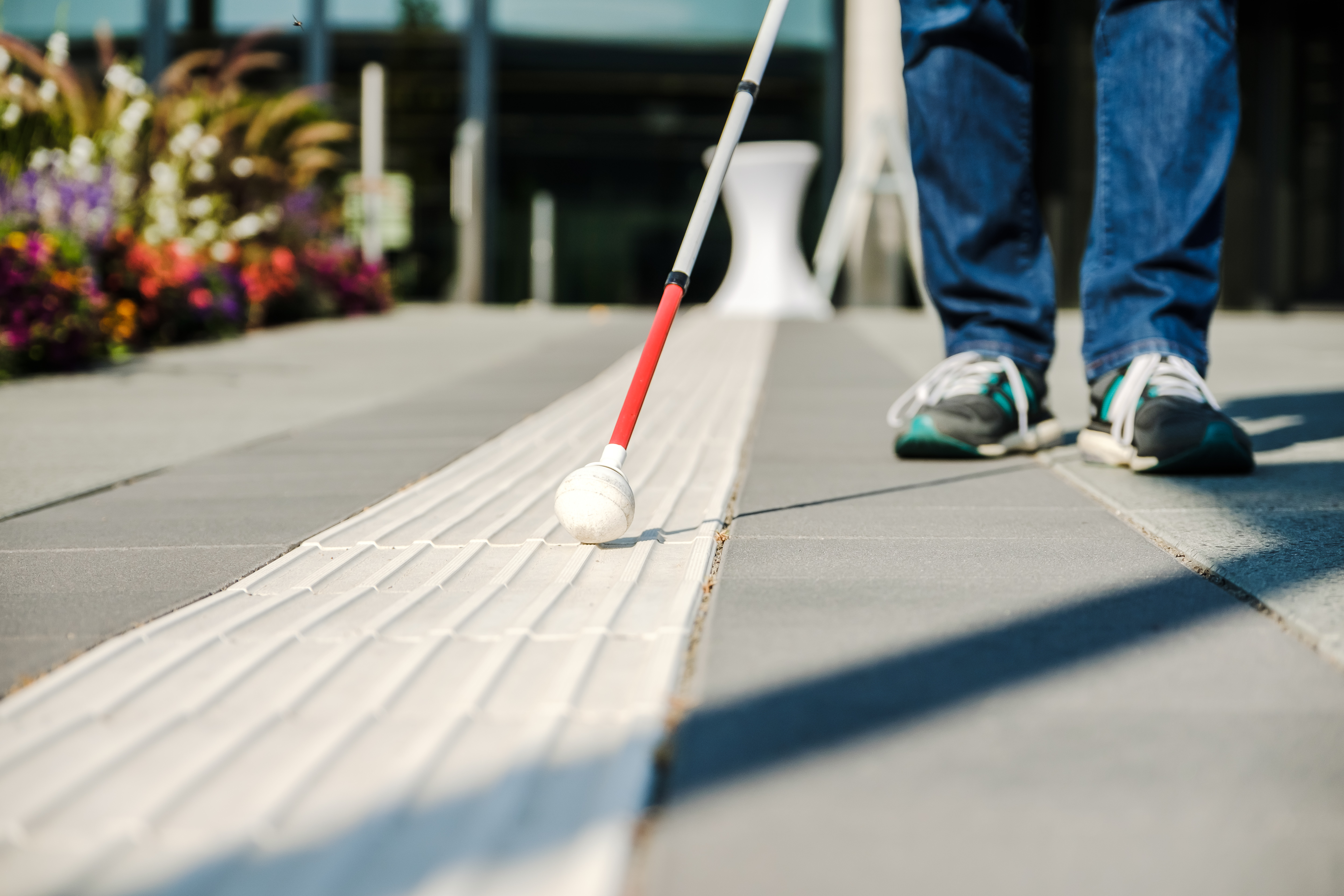 Nahaufnahme von einem Blindenleitsystem auf der Stra&szlig;e, die F&uuml;&szlig;e von einem Menschen sind zu sehen, dieser h&auml;lt einen Blindenstock und geht das Leitsystem entlang.