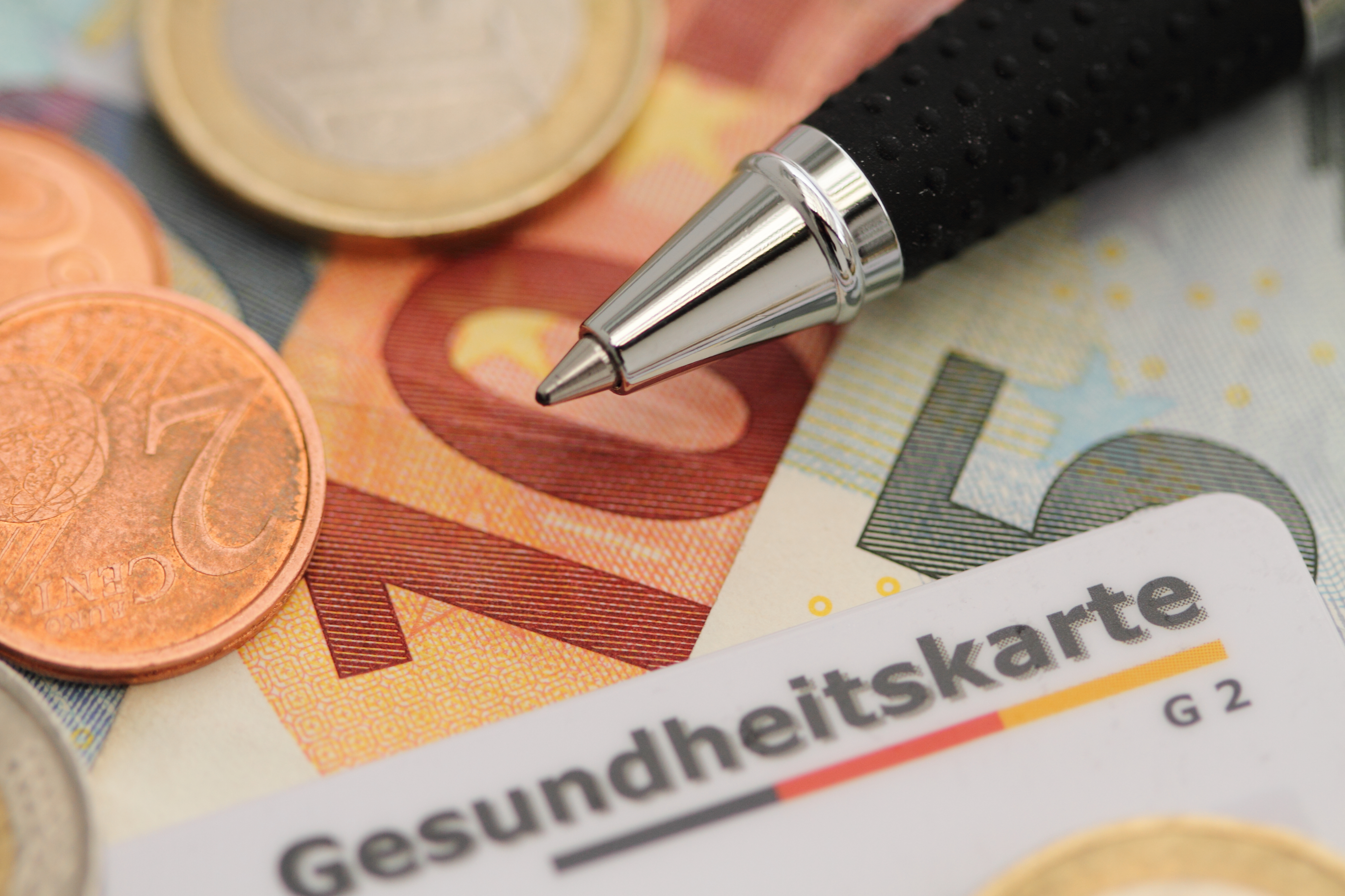 Nahaufnahme von Eurom&uuml;nzen und Scheinen, daneben liegt eine Gesundheitskarte der Krannkenkasse, au&szlig;erdem ein Kugelschreiber.