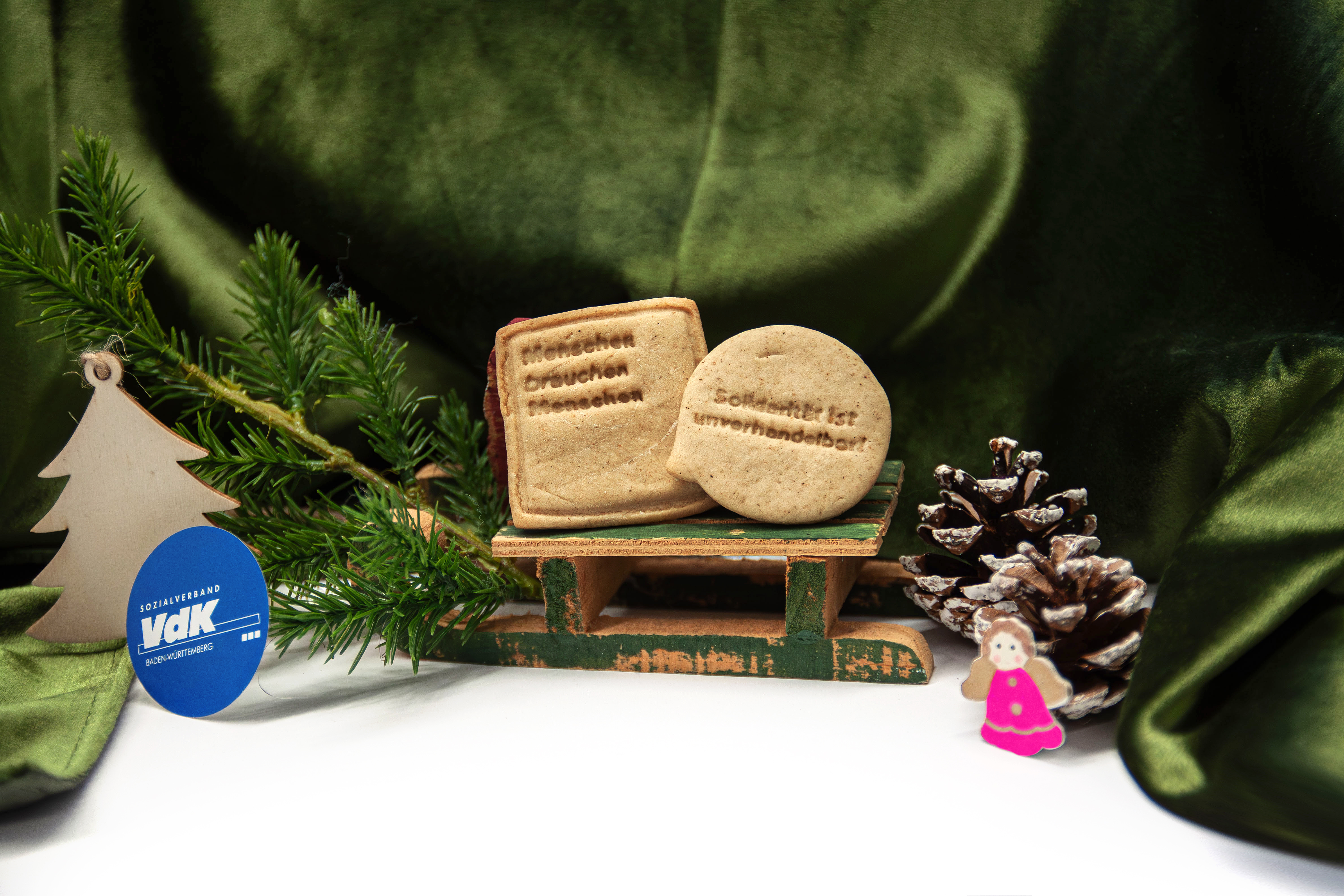 Nahaufnahme eines kleinen Schlittens, auf dem zwei Pl&auml;tzchen stehen. Auf diesen steht geschrieben: "Menschen brauchen Menschen" und "Solidarit&auml;t ist unverhandelbar!". Neben dem Schlitten ist es weihnachtlich dekoriert, mit einer gr&uuml;nen samtigen Decke, einem Tannenzweig sowie Tannenzapfen.