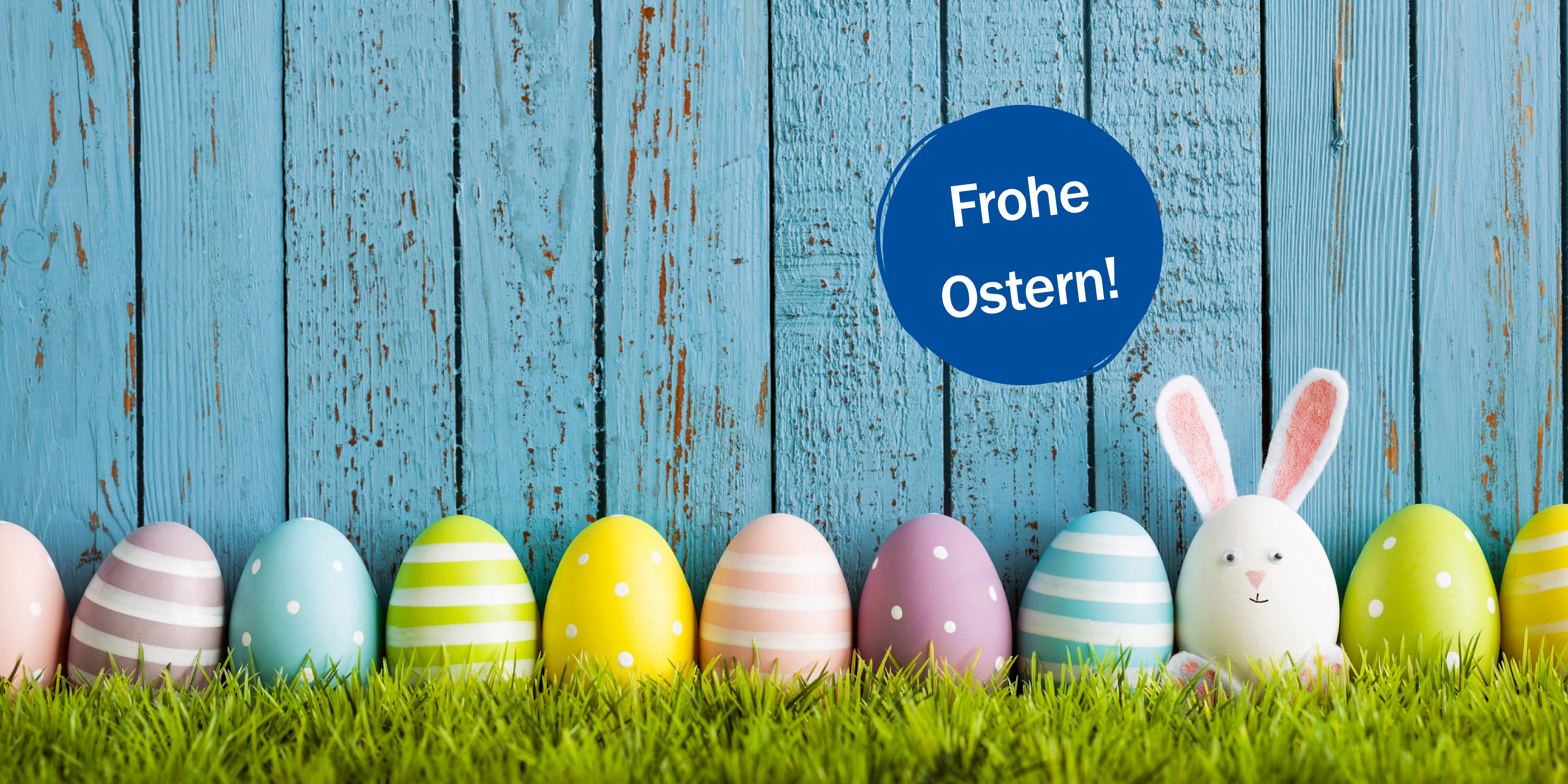 Vor einem h&ouml;lzernen Lattenzaun sind nebeneinander aufgereiht auf einem gr&uuml;nen Rasen viele bunte Ostereier, dazwischen ein wei&szlig;es H&uuml;hnerei mit angeklebten Hasenohren und einem H&auml;schengesicht. Auf einem kreisrunden blauen St&ouml;rer ist die Aufschrift "Frohe Ostern!" zu lesen.