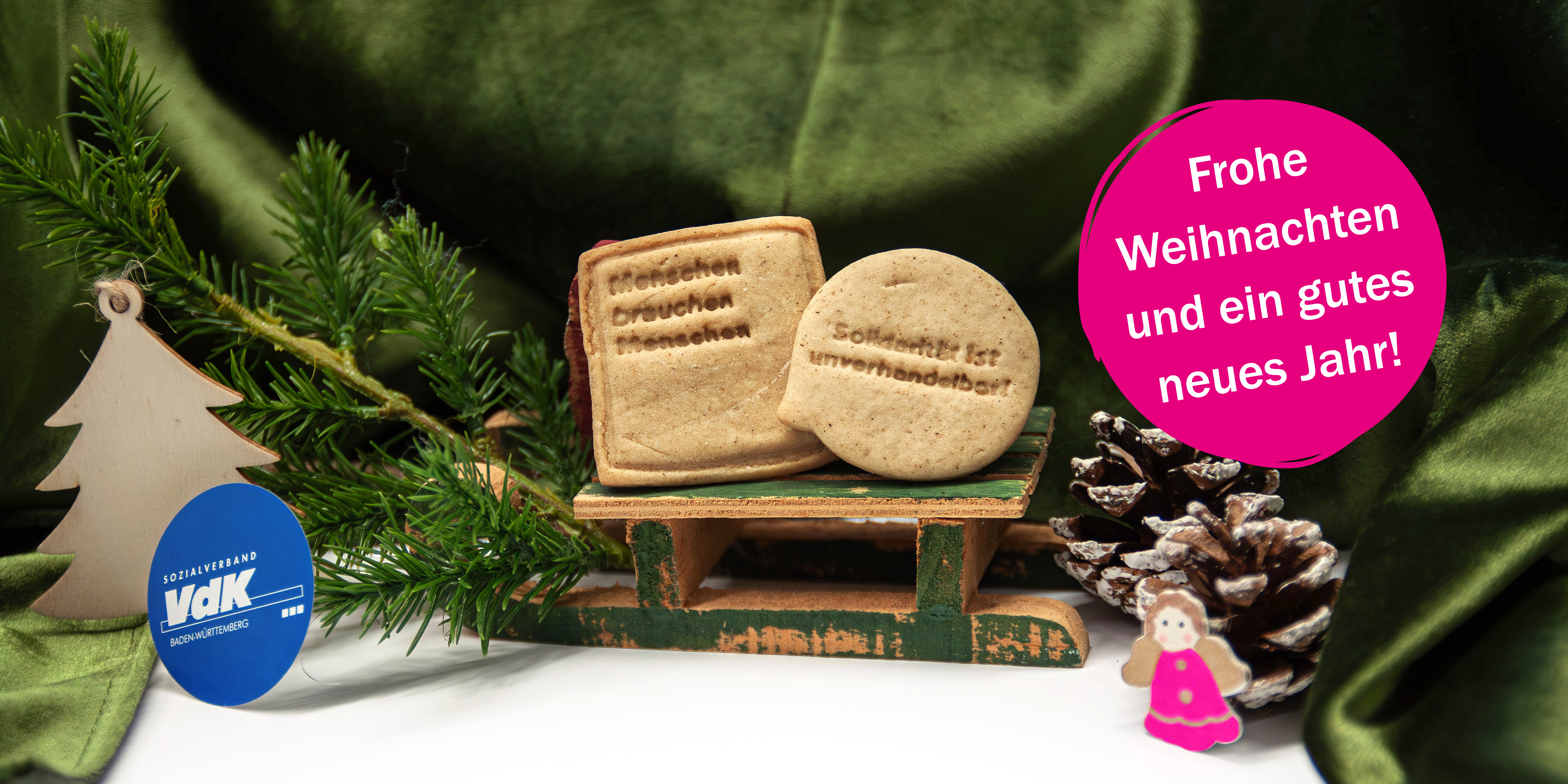 Nahaufnahme eines kleinen Schlittens, auf dem zwei Pl&auml;tzchen stehen. Auf diesen steht geschrieben: "Menschen brauchen Menschen" und "Solidarit&auml;t ist unverhandelbar!". Neben dem Schlitten ist es weihnachtlich dekoriert, mit einer gr&uuml;nen samtigen Decke, einem Tannenzweig sowie Tannenzapfen.