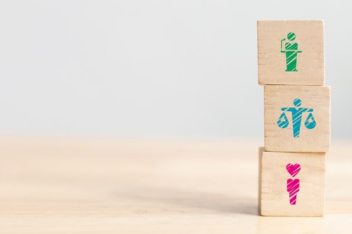 Sozialverband VdK-Baden-Württemberg 3 Säulen Sozialrecht Sozialpolitik und Solidargemeinschaft Drei Holzwürfel stehen auf einem hölzernen Tisch übereinandergestapelt, auf jedem Würfel ist ein anderes Piktogramm zu stehen, eine rosane Figur mit einem Herzen als Kopf, eine türkisfarbene Figur, die zwei Waagen hält und dabei die Arme ausstreckt, und eine grüne Figur die eine Rede an einem Rednerpult hält.