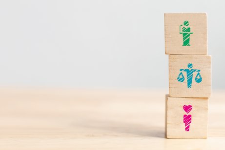 Drei Holzw&uuml;rfel stehen auf einem h&ouml;lzernen Tisch &uuml;bereinandergestapelt, auf jedem W&uuml;rfel ist ein anderes Piktogramm zu stehen, eine rosane Figur mit einem Herzen als Kopf, eine t&uuml;rkisfarbene Figur, die zwei Waagen h&auml;lt und dabei die Arme ausstreckt, und eine gr&uuml;ne Figur die eine Rede an einem Rednerpult h&auml;lt.