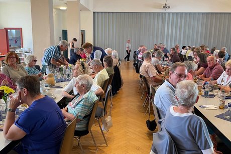 Panorama des Festsaals mit zwei Tischreihen, an denen eine bunt gemischte, fr&ouml;hliche Runde von Menschen sitzt und sich angeregt unterh&auml;lt.