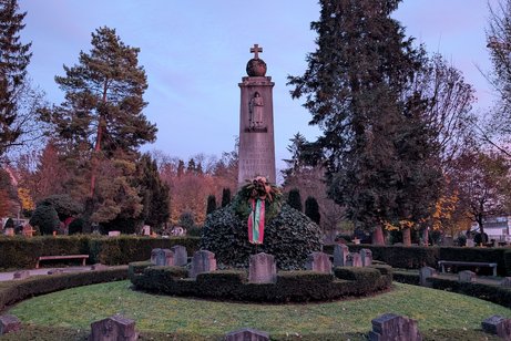 Rechteckige Steinsäule mit einer Kugel und einem Kreuz an der Spitze. Um die Säule herum erstreckt sich eine runde Grünfläche, die von einer niedrigen Hecke umgeben ist. An der Säule lehnt ein großer Kranz.
