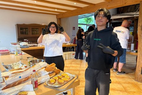 Das Bild zeigt 2 Jugendliche (ein Mädchen und ein Junge), die gerade die Knoblauchbaguette vorbereiten, lachen und in die Kamera das Pease-Zeichen zeigen.