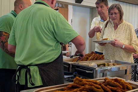 Essensausgabe. Von hinten stehen zwei Männer im grünen T-Shirt hinter einem Tisch mit Warmhaltebehältern. Davor eine Menschenschlange. Ganz vorne hält eine Frau einen Teller mit Essen, neben ihr zeigt ein Mann auf eine Essensbox. Im Vordergrund ist ein Warmhaltebehälter zu sehen, gefüllt mit zahlreichen panierten Schnitzeln.