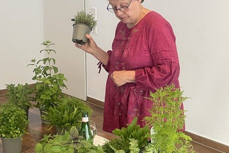 Eine Frau mit Kurzhaarschnitt und Brille in einem weinroten Kleid steht an einem Holztisch und h&auml;lt einen Topf mit Kr&auml;utern in der Hand. Vor ihr stehen mehrere weitere Kr&auml;utert&ouml;pfe, eine Flasche Wasser und ein Glas. Im Hintergrund ist eine wei&szlig;e T&uuml;r mit einem gr&uuml;nen Rettungszeichen angebracht.