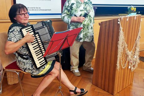 Unsere Musik: Anita Akkordeon, Inge Gesang Anita sitzend am Akkordeon vorne links. Inge mit Mikrofon in der Hand neben dem Rednerpult.
