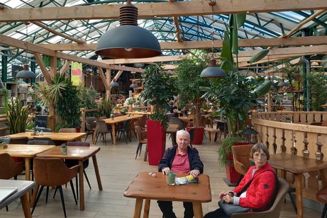 Kaffeebereich in einem gro&szlig;en Gew&auml;chshaus mit mehreren Holztischen und St&uuml;hlen, an einem Tisch sitzen zwei &auml;ltere Damen, dazwischen stehen mehrere Palmen in Pflanzk&uuml;beln.