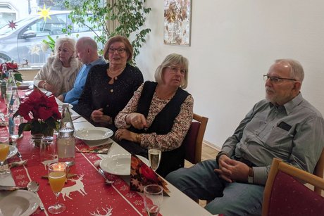 Drei Damen und zwei Herren sitzen an einem Tisch, einige unterhalten sich, andere blicken in Richtung Kamera.