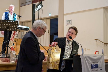 Ein Herr in schickem Anzug überreicht einem anderen Mann im legeren Anzug ein Weinpräsent.