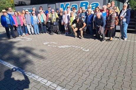 Eine Gruppe von etwa 30 Personen &ndash; &uuml;berwiegend &auml;ltere Menschen &ndash; steht im Freien vor einem Transparent mit blauer Beschriftung &bdquo;MaBa&ldquo;. Die Gruppe besteht aus M&auml;nnern und Frauen unterschiedlichen Alters, einige tragen H&uuml;te oder Sonnenbrillen, einer nutzt einen Rollator. Sie blicken in Richtung Kamera, einige l&auml;cheln. Im Vordergrund ist ein gepflasterter Platz mit Schattenspiegeln von B&auml;umen zu sehen. Die Szene wirkt fr&ouml;hlich und gemeinschaftlich.