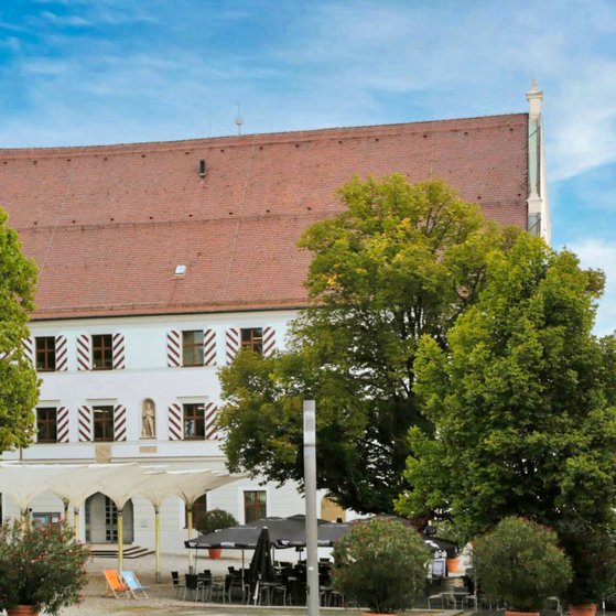 Ein weißes, dreistöckiges Gebäude mit rot-weiß gestreiften Fensterläden. Im Vordergrund stehen schattenspendende Bäume sowie gemütliche Sitzgelegenheiten.