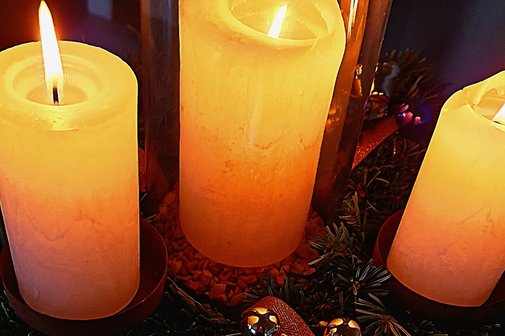 Man sieht drei brennende Kerzen nebeneinander und darunter Tannenzweige sowie etwas Rosa Band und kleine goldene Kugeln.