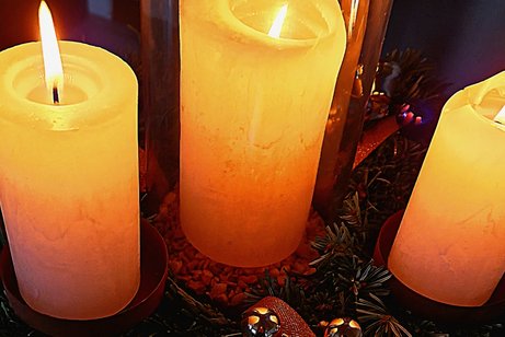 Man sieht drei brennende Kerzen nebeneinander und darunter Tannenzweige sowie etwas Rosa Band und kleine goldene Kugeln.