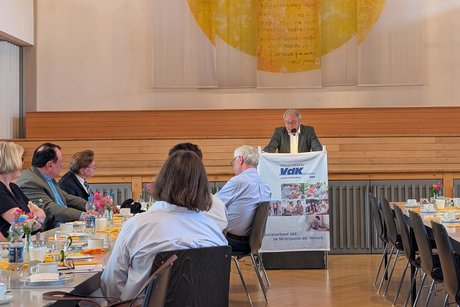 Menschen sitzen an einem Tisch und hören einem älteren Herrn am Rednerpult aufmerksam zu.