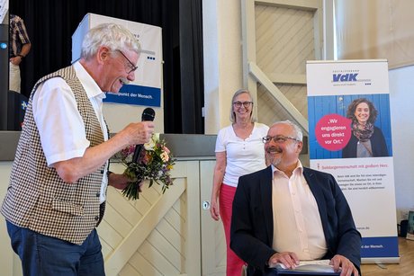 Ein Mann im weißen Kurzarmhemd überreicht einem Rollstuhlfahrer einen Blumenstrauß.