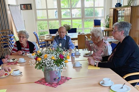 Drei Damen und ein Herr sitzen bei Kaffee und Kuchen an einem Tisch und unterhalten sich angeregt; in der Mitte des Tisches steht ein prächtiger Blumenstrauß.