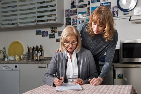 Eine bebrillte, ältere Frau Tisch sitzend füllt ein Formular aus, eine jüngere Frau hinter ihr stehend schaut ihr über die Schulter, eine Hand hat die stehende Frau der sitzenden Frau auf die rechte Schulter gelegt .